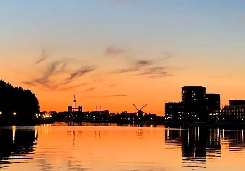 Op de foto ziet u Gouda bij avond. De foto is genomen vanaf het Gouwekanaal. Op de achtergrond ziet u de spoorbrug, een windmolen, een molen, huizen en kantoren. De lucht is oranje gekleurd. 