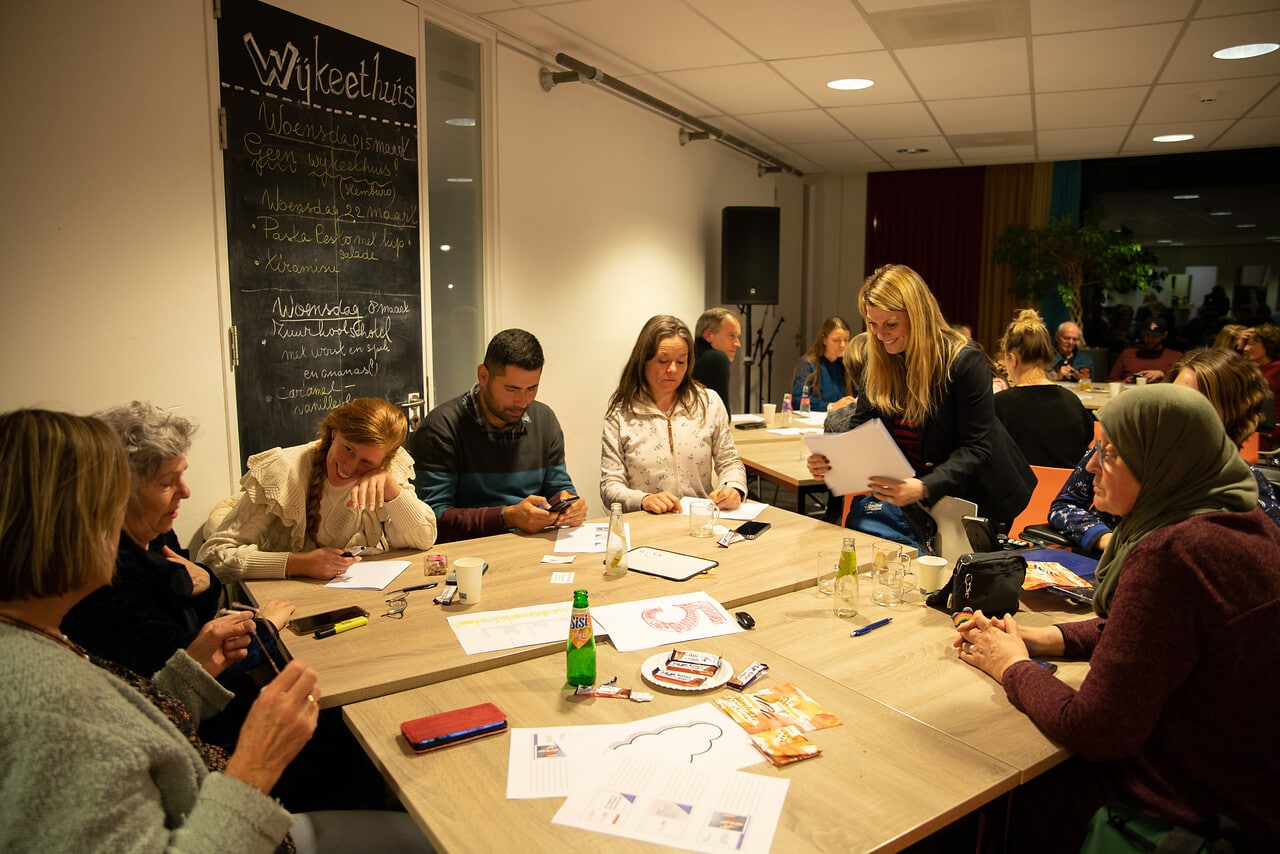 Op de foto zitten verschillende mensen bij elkaar aan tafel in een wijkcentrum. Een vrouw deelt papier uit en mensen maken aantekeningen.