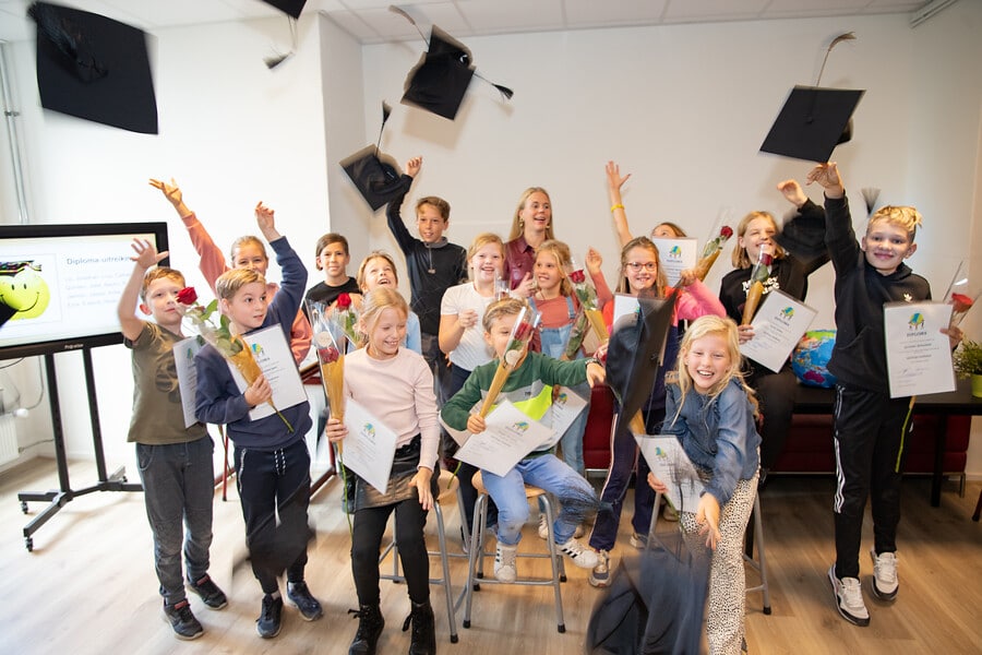 Op de foto staat een groep basisschoolkinderen, die afstudeerhoeden de lucht in gooien. Ze hebben ieder een certificaat en een rode roos in hun hand. Ook staat wethouder Anna van Popering-Kalkman op de foto.